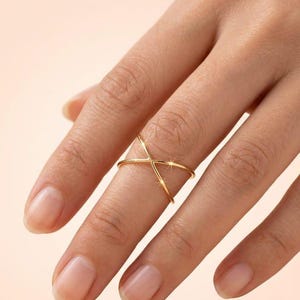May include: A polished gold ring featuring a slender, minimalist band that crosses over itself in an X-shape design. The metal has a smooth, reflective surface that catches the light, creating a subtle sparkle at the intersection point.