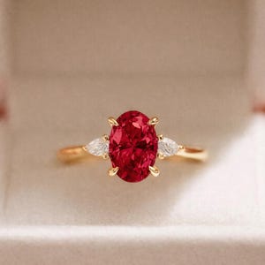 Könnte beinhalten: Ein goldener Ring mit einem großen, oval geschliffenen Rubin als Mittelstein. Zwei birnenförmige, weiße Edelsteine flankieren den Rubin. Der Ring ist in einem goldenen Band gefasst und wird in einer weißen Schmuckschatulle präsentiert.