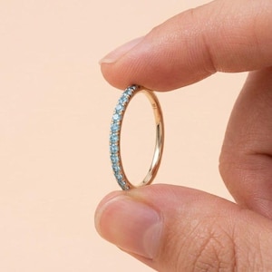 Op de afbeelding: Een gouden bandring met een rij kleine, lichtblauwe edelstenen. De ring wordt tussen twee vingers gehouden tegen een perzikkleurige achtergrond. De ring is ongeveer 0,25 cm breed.
