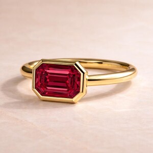 Pode incluir: Um anel de ouro com uma pedra preciosa vermelha escura retangular e facetada. A pedra preciosa &eacute; engastada em uma moldura de ouro, e a banda &eacute; de ouro liso e polido. O anel &eacute; exibido em uma superf&iacute;cie rosa claro.