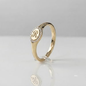 Pode incluir: Um anel de ouro com uma face oval apresentando a letra R gravada ao centro, rodeada por uma borda de pequenas pedras claras. O aro é liso e polido, com o texto 14K claramente visível na parte interna da haste metálica do anel.