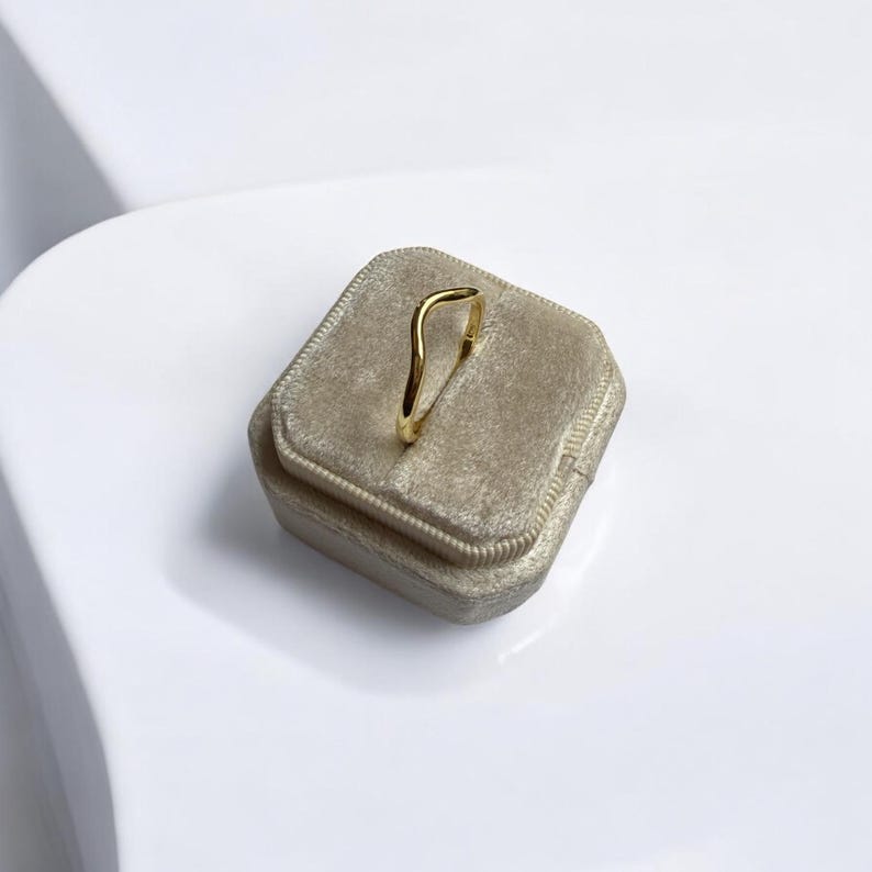 K&ouml;nnte beinhalten: Ein goldfarbener Ring mit wellenf&ouml;rmigem Design, auf einer beigefarbenen Samt-Ringbox. Der Ring hat eine glatte, polierte Oberfl&auml;che und einen minimalistischen Stil. Die Ringbox ist quadratisch mit einer weichen, strukturierten Oberfl&auml;che.