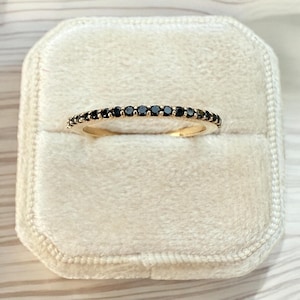 May include: A gold band ring with a row of small, black gemstones. The ring is set against a cream-colored, velvet ring box. The ring is a delicate piece of jewelry.