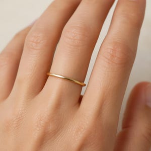 Op de afbeelding: Een dunne, goudkleurige ring wordt aan een vinger gedragen. De ring heeft een eenvoudig, minimalistisch ontwerp en een gepolijste afwerking. De ring lijkt van metaal te zijn en reflecteert licht.