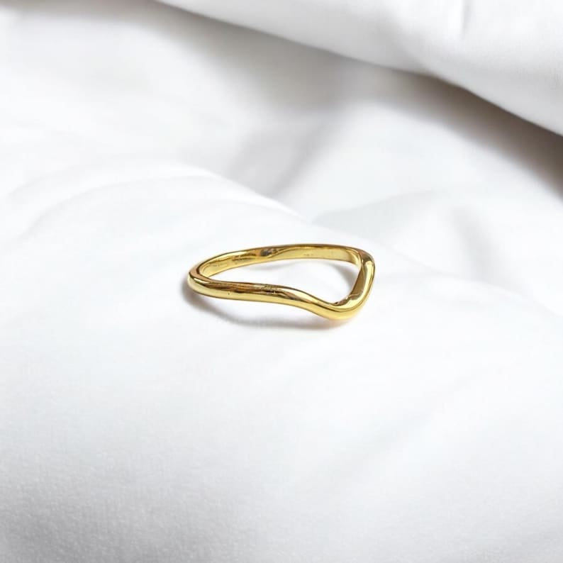 K&ouml;nnte beinhalten: Ein goldfarbener Ring mit einem wellenf&ouml;rmigen, V-f&ouml;rmigen Design. Der Ring ist vor einem wei&szlig;en Hintergrund platziert, der seine glatte, reflektierende Oberfl&auml;che hervorhebt. Die einzigartige Form des Rings verleiht ihm einen modernen Touch.