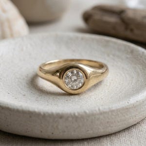 Op de afbeelding: Een gouden ring met een enkele ronde, briljant geslepen heldere edelsteen in een gladde, gepolijste zetting. De ringband is van massief goud met een taps toelopend ontwerp dat breder wordt naar het midden, voor een strak en rond profiel.