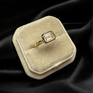 May include: A gold ring featuring a clear emerald-cut gemstone set in a bezel setting. The ring is presented in a white velvet ring box.