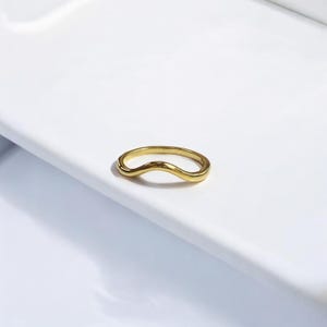 K&ouml;nnte beinhalten: Ein goldfarbener Ring mit wellenf&ouml;rmigem Design. Der Ring besteht aus einem gl&auml;nzenden, reflektierenden Metall und liegt auf einer wei&szlig;en Oberfl&auml;che. Das Design des Rings ist eine durchgehende, flie&szlig;ende Wellenform.