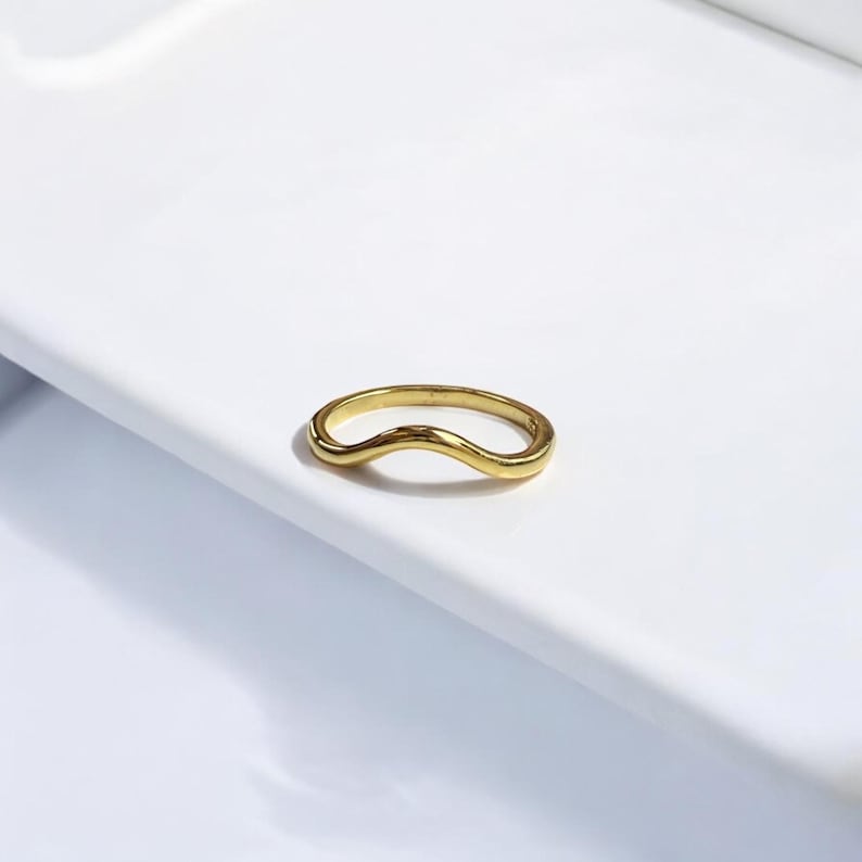 K&ouml;nnte beinhalten: Ein goldfarbener Ring mit wellenf&ouml;rmigem Design. Der Ring besteht aus einem gl&auml;nzenden, reflektierenden Metall und liegt auf einer wei&szlig;en Oberfl&auml;che. Das Design des Rings ist eine durchgehende, flie&szlig;ende Wellenform.