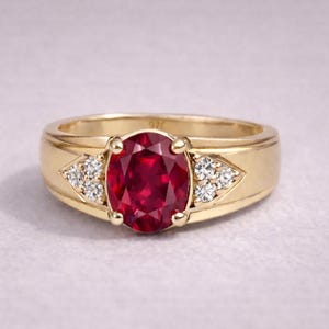 May include: A gold ring with a large oval ruby gemstone. The ring has a wide band and is decorated with small diamonds on either side of the ruby. The ring is set against a light background.