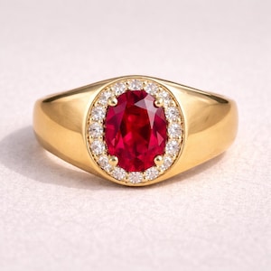 Peut inclure: Une bague en or ornée d'une grande pierre rouge vif de forme ovale, entourée d'un halo de petites pierres claires. La bague présente un anneau lisse et poli et un design classique.