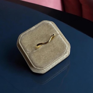 K&ouml;nnte beinhalten: Ein goldfarbener Ring mit wellenf&ouml;rmigem Design liegt in einer beigefarbenen Samt-Ringbox. Der Ring hat eine glatte, polierte Oberfl&auml;che und eine geschwungene Form. Die Ringbox ist rechteckig und hat eine weiche, pl&uuml;schige Textur. Der Hintergrund ist eine dunkelblaue Oberfl&auml;che.