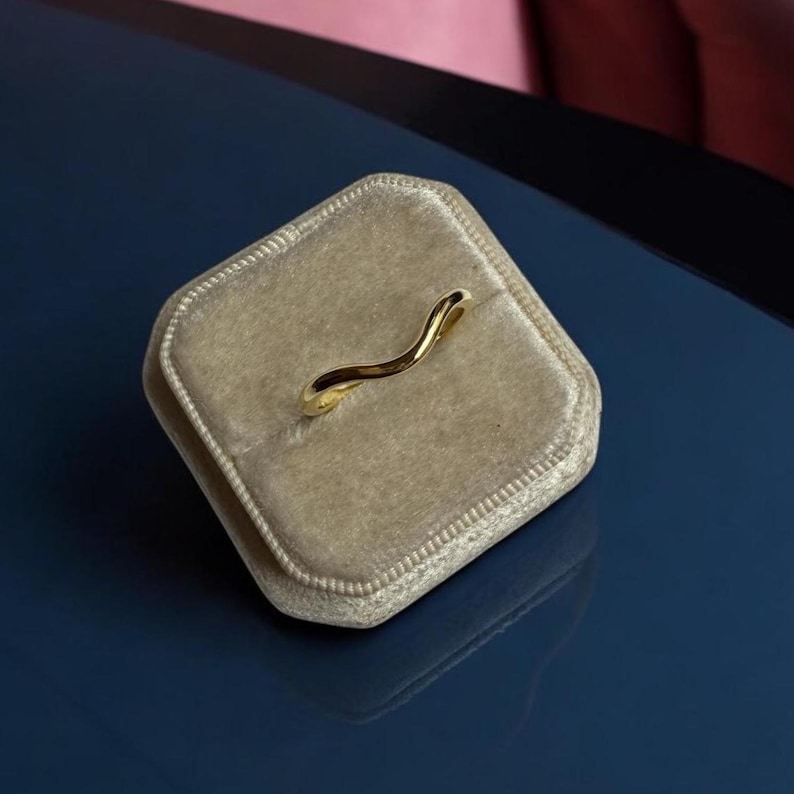 K&ouml;nnte beinhalten: Ein goldfarbener Ring mit wellenf&ouml;rmigem Design liegt in einer beigefarbenen Samt-Ringbox. Der Ring hat eine glatte, polierte Oberfl&auml;che und eine geschwungene Form. Die Ringbox ist rechteckig und hat eine weiche, pl&uuml;schige Textur. Der Hintergrund ist eine dunkelblaue Oberfl&auml;che.