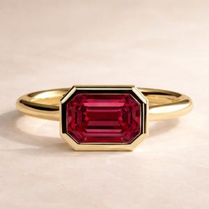 May include: A gold ring featuring a rectangular, deep red gemstone. The gemstone is set within a gold bezel, and the band is a smooth, polished gold. The ring is displayed on a light-colored surface, highlighting its elegant design.