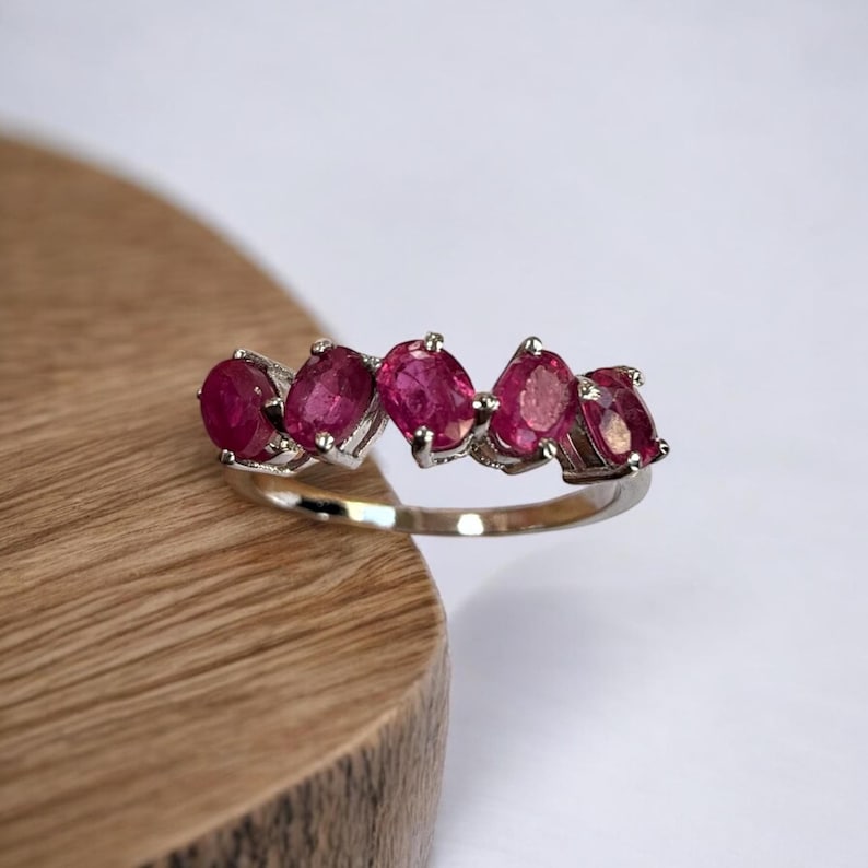 Peut inclure: Une bague en argent avec cinq pierres pr&eacute;cieuses rubis de forme ovale serties en rang&eacute;e. La bague est pos&eacute;e sur une surface en bois.