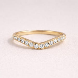 Puede incluir: Un anillo dorado curvado con una fila de pequeñas piedras preciosas transparentes en la parte superior. El anillo tiene un diseño delicado, de inspiración vintage, perfecto para combinar con otros anillos. La banda es lisa y pulida.