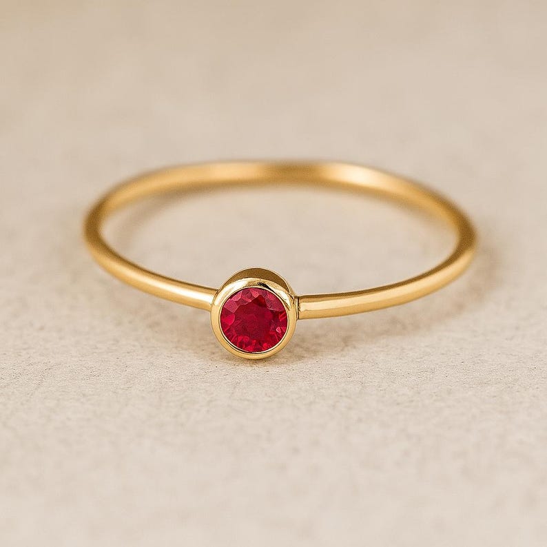 May include: A gold ring with a round, bright red gemstone. The ring has a thin band and a simple, elegant design. The gemstone is set in a bezel setting, and the ring is likely made of gold.