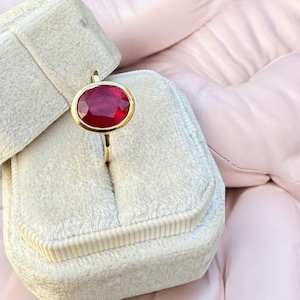 May include: A gold ring with a large, oval-shaped, deep red gemstone. The ring is set in a gold bezel and is displayed in a light gray velvet ring box against a pink background.