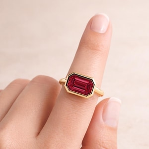 Pode incluir: Um anel de ouro com uma pedra preciosa retangular vermelha escura. A pedra preciosa &eacute; colocada em uma moldura de ouro, criando um contraste marcante. O anel &eacute; mostrado em um dedo, destacando seu design e cor.