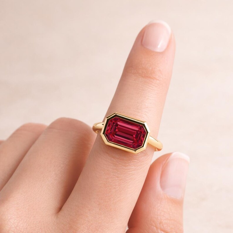 Pode incluir: Um anel de ouro com uma pedra preciosa retangular vermelha escura. A pedra preciosa &eacute; colocada em uma moldura de ouro, criando um contraste marcante. O anel &eacute; mostrado em um dedo, destacando seu design e cor.