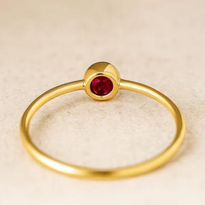 May include: A gold ring with a round, bright red gemstone. The ring has a thin, polished band and a simple, elegant design. The gemstone is set in a gold bezel setting, and the ring is shown against a neutral background.
