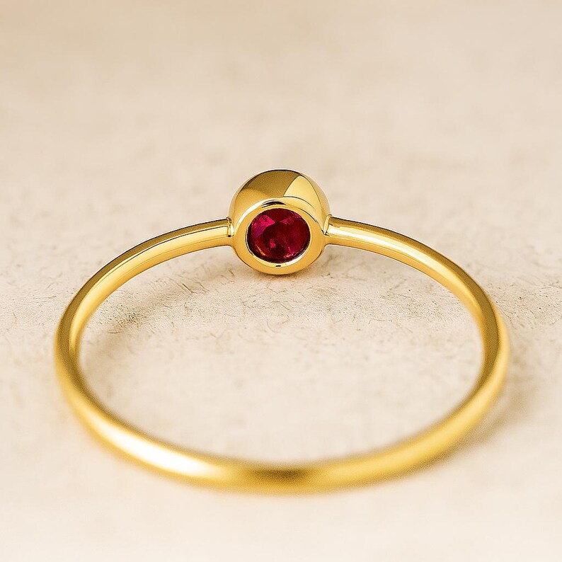 May include: A gold ring with a round, bright red gemstone. The ring has a thin, polished band and a simple, elegant design. The gemstone is set in a gold bezel setting, and the ring is shown against a neutral background.