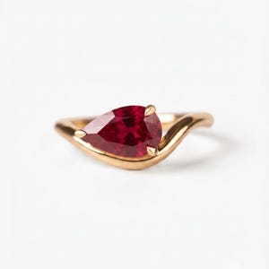 Pode incluir: Um anel de ouro com uma pedra preciosa vermelho-escura em forma de pera, fixada por garras. A banda tem um design liso, curvo e fluido que envolve a base da pedra, criando uma silhueta metálica polida e contínua.