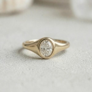 May include: A gold signet ring featuring a central oval-shaped clear gemstone set within a smooth, polished metal band. The ring has a tapered design that widens towards the centre, showcasing a bright, reflective finish and a faceted stone.