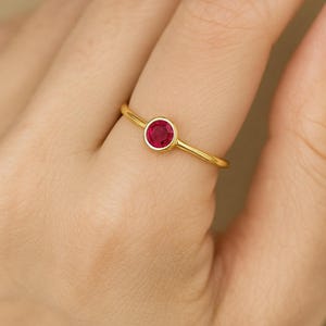 May include: A gold ring with a round, bright red gemstone. The ring has a thin band and a bezel setting that holds the gemstone securely. The ring is shown on a person's finger.