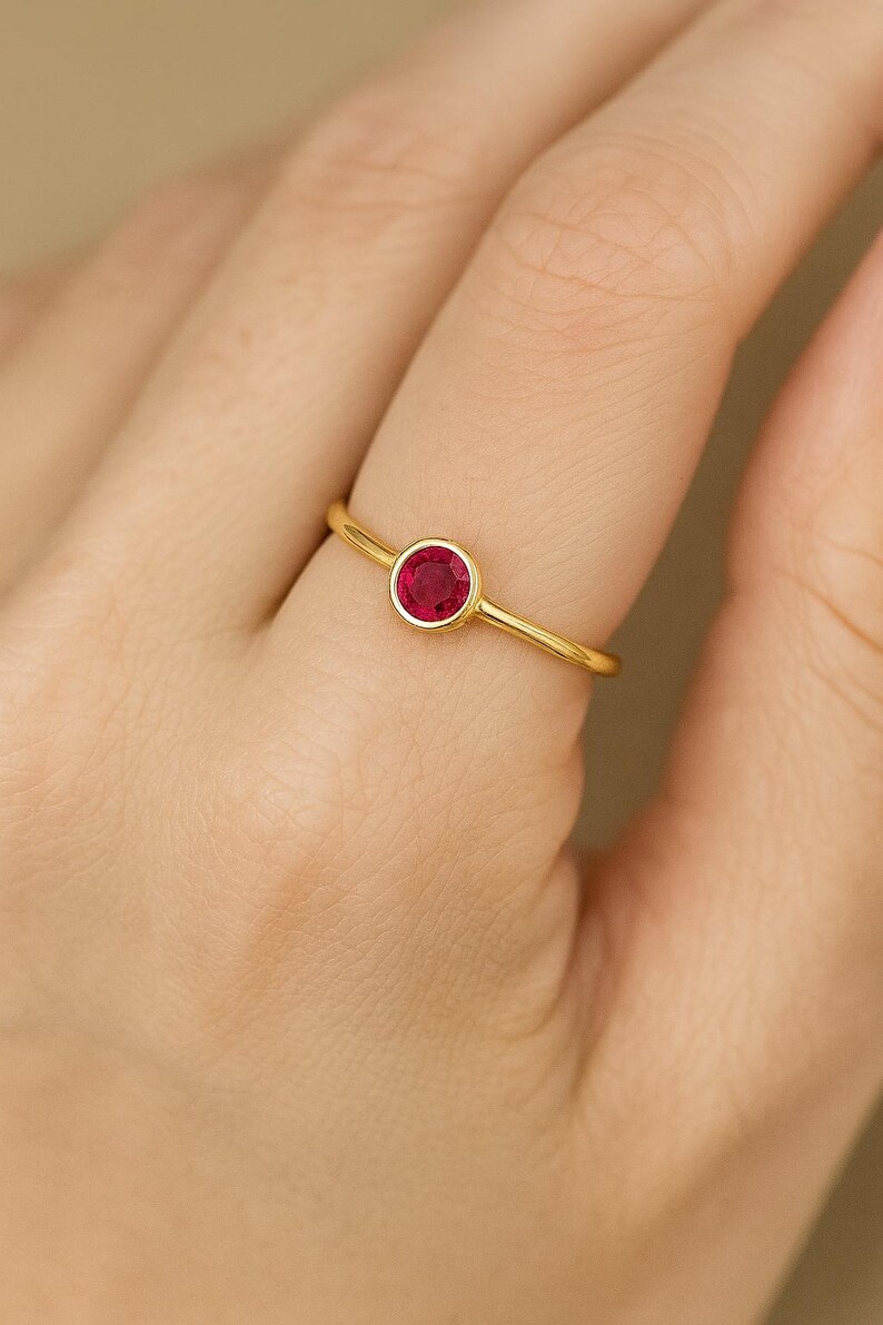 May include: A gold ring with a round, bright red gemstone. The ring has a thin band and a bezel setting that holds the gemstone securely. The ring is shown on a person's finger.
