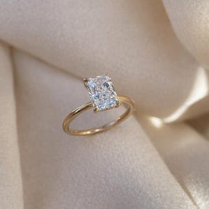 以下が含まれることがあります： 長方形カットのダイヤモンドが輝くゴールドリング。 ダイヤモンドは明るく輝いています。 リングは細いゴールドバンドで、クラシックで洗練された美しさを提供します。 リングは柔らかくニュートラルな色の生地の上に展示されています。