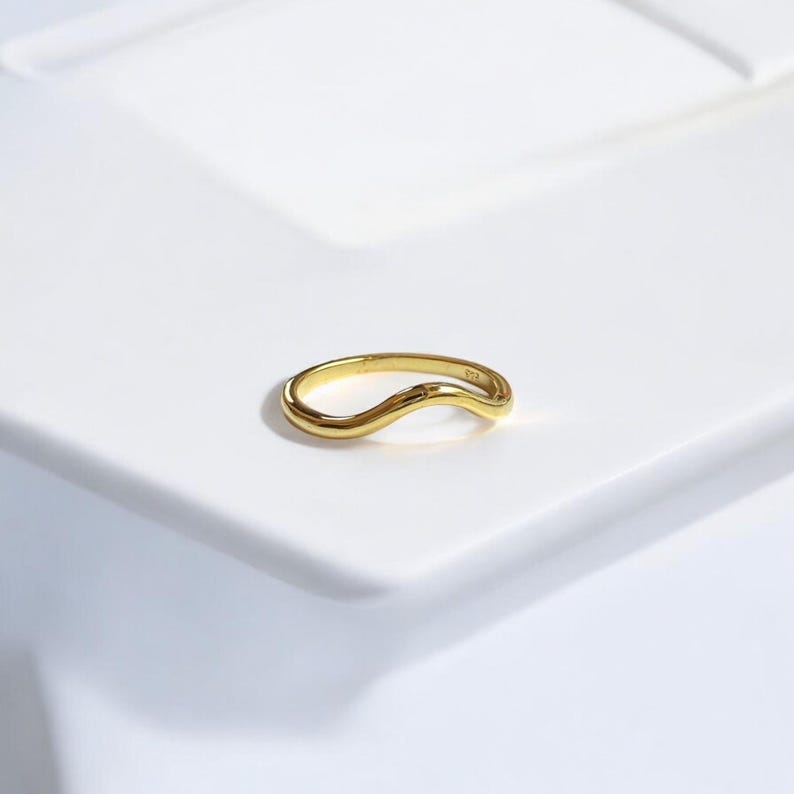 K&ouml;nnte beinhalten: Ein goldfarbener Ring mit wellenf&ouml;rmigem Design. Der Ring ist vor einem wei&szlig;en Hintergrund platziert, der seine glatte, geschwungene Form hervorhebt. Das Design des Rings ist schlicht und elegant.