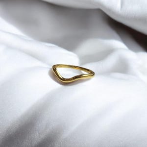 K&ouml;nnte beinhalten: Ein goldfarbener Ring mit einem wellenf&ouml;rmigen, organischen Design. Der Ring ist vor einem wei&szlig;en, strukturierten Hintergrund platziert, der seinen Glanz und seine Form hervorhebt. Die Oberfl&auml;che des Rings reflektiert das Licht und betont seine metallische Ausf&uuml;hrung.