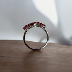 Peut inclure: Une bague en argent avec cinq pierres pr&eacute;cieuses rouges serties en rang&eacute;e. La bague est pos&eacute;e sur une surface en bois.