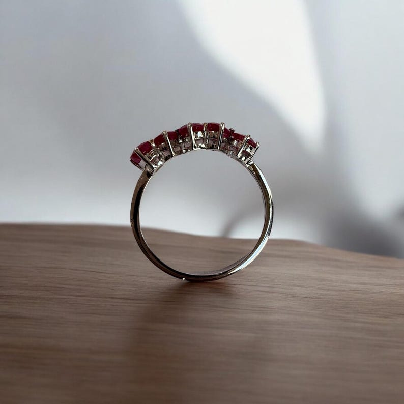 Peut inclure: Une bague en argent avec cinq pierres pr&eacute;cieuses rouges serties en rang&eacute;e. La bague est pos&eacute;e sur une surface en bois.