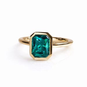 Op de afbeelding: Een gouden ring met een rechthoekige, smaragdgeslepen, teal-kleurige edelsteen. De edelsteen is gezet in een gouden bezel en de band is van effen goud. De ring wordt getoond tegen een witte achtergrond.