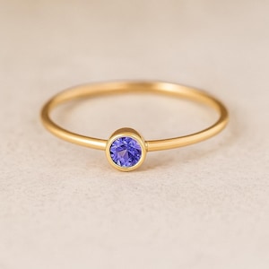 May include: A delicate gold ring featuring a round, vibrant blue gemstone. The ring has a thin band and a simple, elegant design. The gemstone is set in a bezel setting, enhancing its color and brilliance. This piece is suitable for everyday wear.