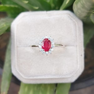 Puede incluir: Un anillo de plata con una piedra preciosa ovalada de rubí rojo rodeada por un halo de piedras blancas más pequeñas.