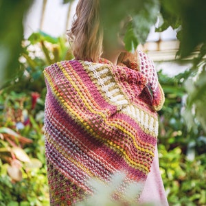 Könnte beinhalten: Ein mehrfarbiger Strickschal in Rot-, Rosa-, Gelb- und Weißtönen. Der Schal hat ein strukturiertes Muster mit diagonalen Streifen und geometrischen Designs. Die Person, die den Schal trägt, befindet sich in einem Garten.