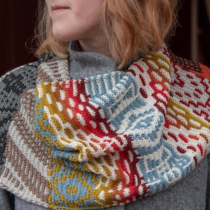Puede incluir: Un cuello tejido colorido con patrones geométricos en tonos grises, marrones, azules, amarillos, rojos y naranjas. El cuello se lleva alrededor del cuello y los hombros, mostrando una variedad de texturas y diseños.
