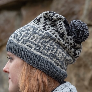 Puede incluir: Un gorro de punto gris, negro y crema con un diseño geométrico y un pompón. El gorro lo lleva una persona con cabello castaño rojizo, con una pared de piedra al fondo. Un accesorio de invierno.