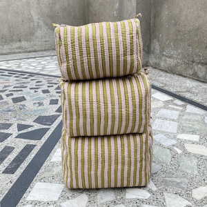 May include: Three stacked square cushions. The cushions are olive green with vertical pink stripes. They are made from a textured fabric and have a simple design. The cushions are stacked on top of each other.