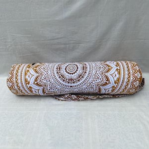 May include: A cylindrical yoga mat bag with a white and gold mandala pattern. The bag has a shoulder strap and a zipper closure. The design features intricate, symmetrical patterns in white against a gold background. The bag is designed to carry a rolled-up yoga mat.