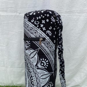 May include: Black and white yoga mat bag with a zipper closure and a shoulder strap. The bag features a detailed, symmetrical pattern with floral and geometric designs. The bag is cylindrical and appears to be made of fabric.