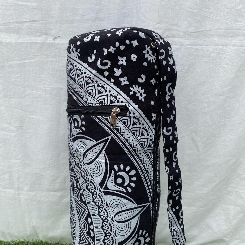 May include: Black and white yoga mat bag with a zipper closure and a shoulder strap. The bag features a detailed, symmetrical pattern with floral and geometric designs. The bag is cylindrical and appears to be made of fabric.