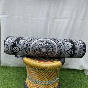 May include: A black and white cylindrical yoga bolster with a detailed mandala design. The bolster is resting on a small, round stool with a woven bamboo frame and a black top. The background is a white sheet.