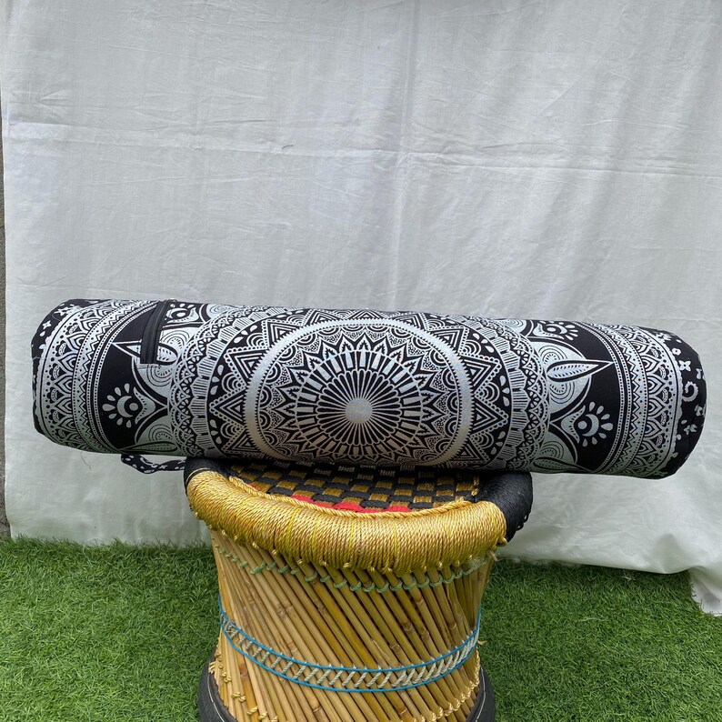 May include: A black and white cylindrical yoga bolster with a detailed mandala design. The bolster is resting on a small, round stool with a woven bamboo frame and a black top. The background is a white sheet.