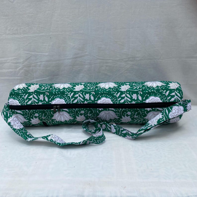 K&ouml;nnte beinhalten: Eine gr&uuml;ne Yogamatten-Tasche mit wei&szlig;em Blumenmuster. Die zylindrische Tasche hat einen schwarzen Rei&szlig;verschluss und einen passenden Schultergurt. Die Tasche ist f&uuml;r den Transport einer Yogamatte und anderem Zubeh&ouml;r konzipiert.