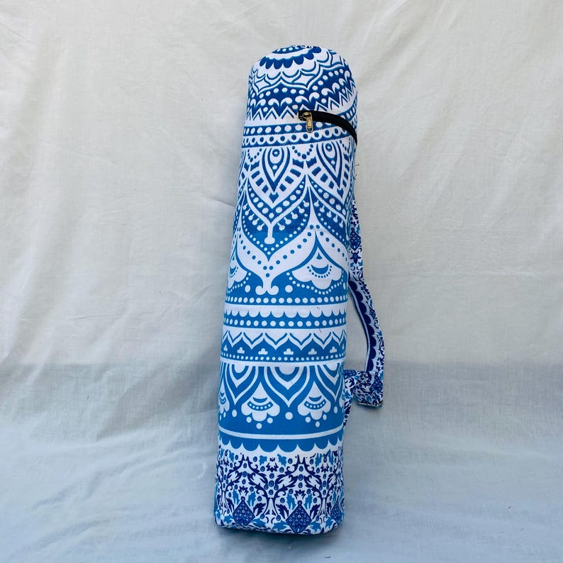 May include: A blue and white yoga mat bag with a decorative mandala pattern. The cylindrical bag has a zipper closure and a shoulder strap. The bag is made of fabric and is designed to carry a yoga mat.
