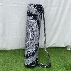 May include: Black and white yoga mat bag with a detailed mandala design. The cylindrical bag has a zipper pocket and a matching strap for carrying. The bag is made of fabric and is designed to hold a yoga mat.